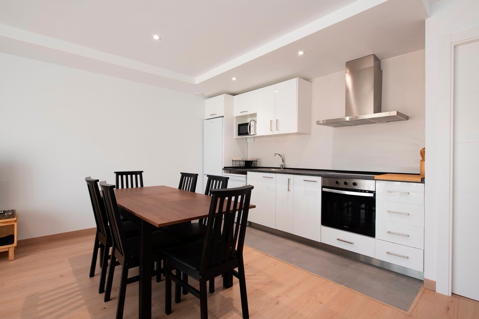 Cocina y comedor modernos con armarios blancos, sillas negras y mesa de madera.