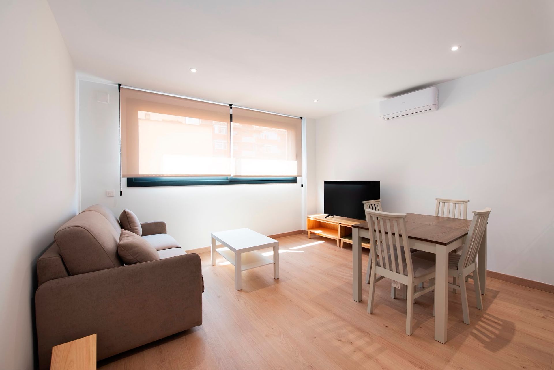 Sala de estar luminosa con sofá marrón, mesa de comedor, televisor y persianas grandes.