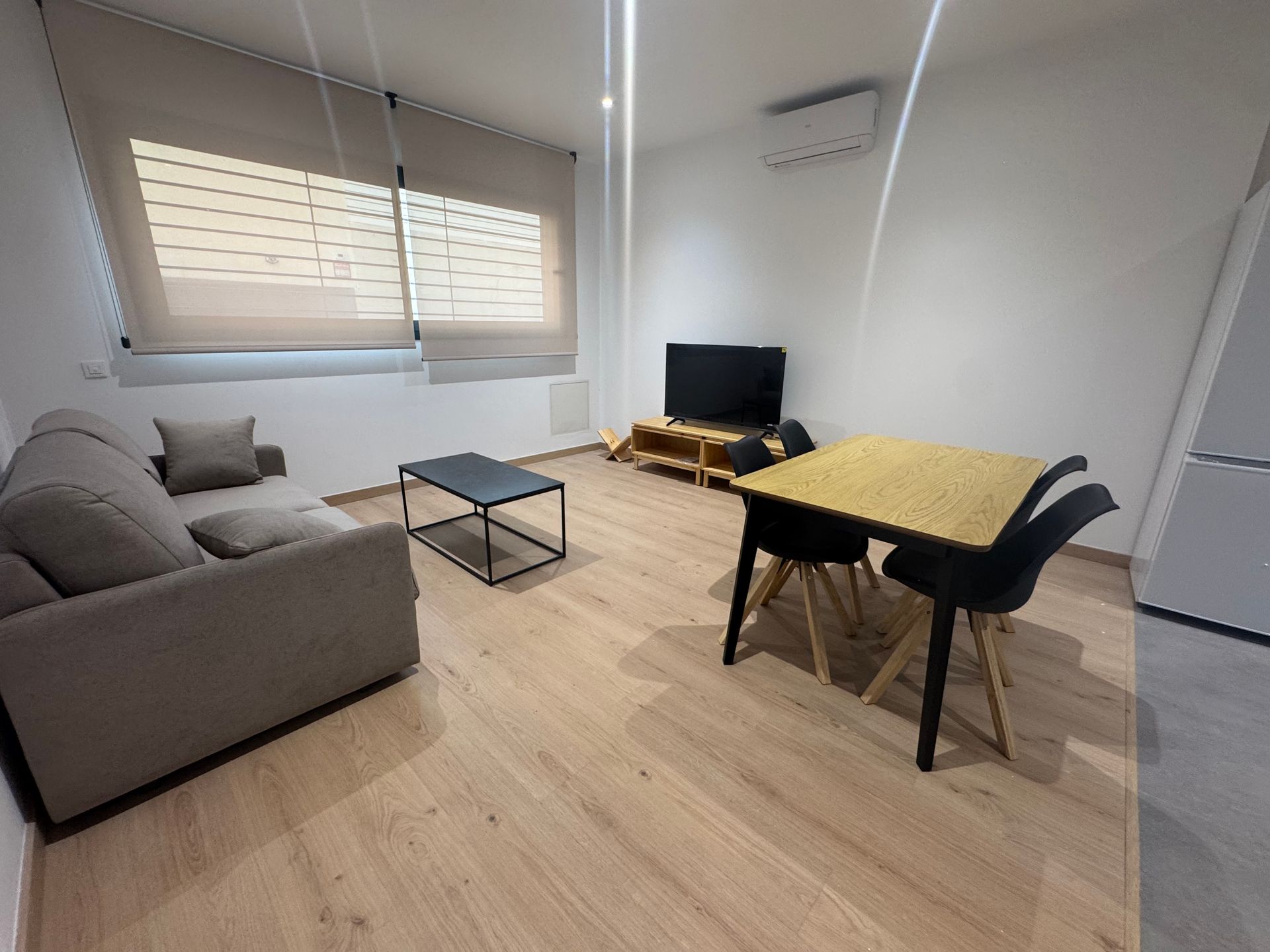 Sala de estar con sofá gris, mesa de centro, mesa de comedor y televisor junto a una ventana con persianas.