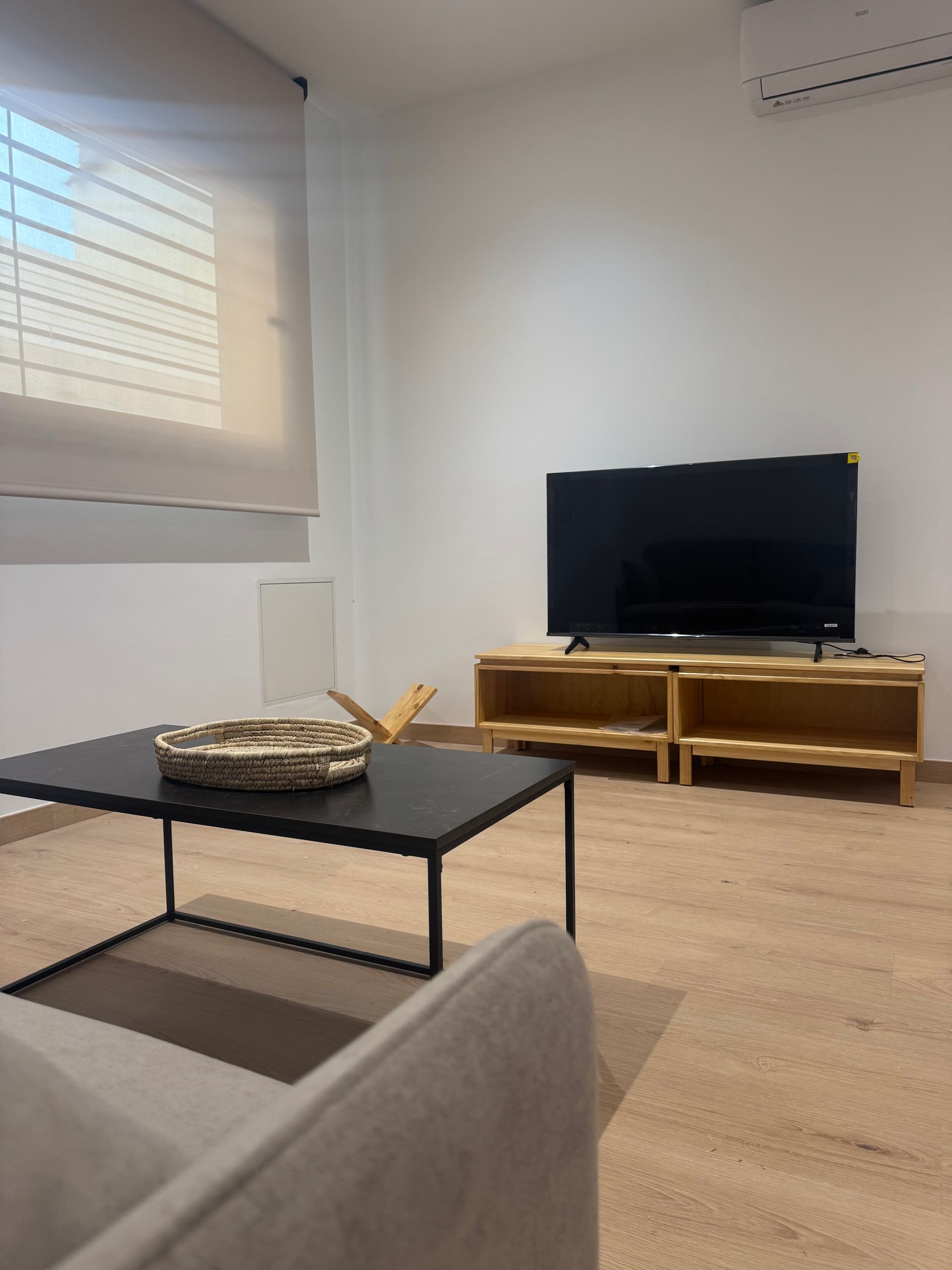 Sala de estar moderna con sofá gris, mesa de centro negra, mueble para TV y persianas grandes.