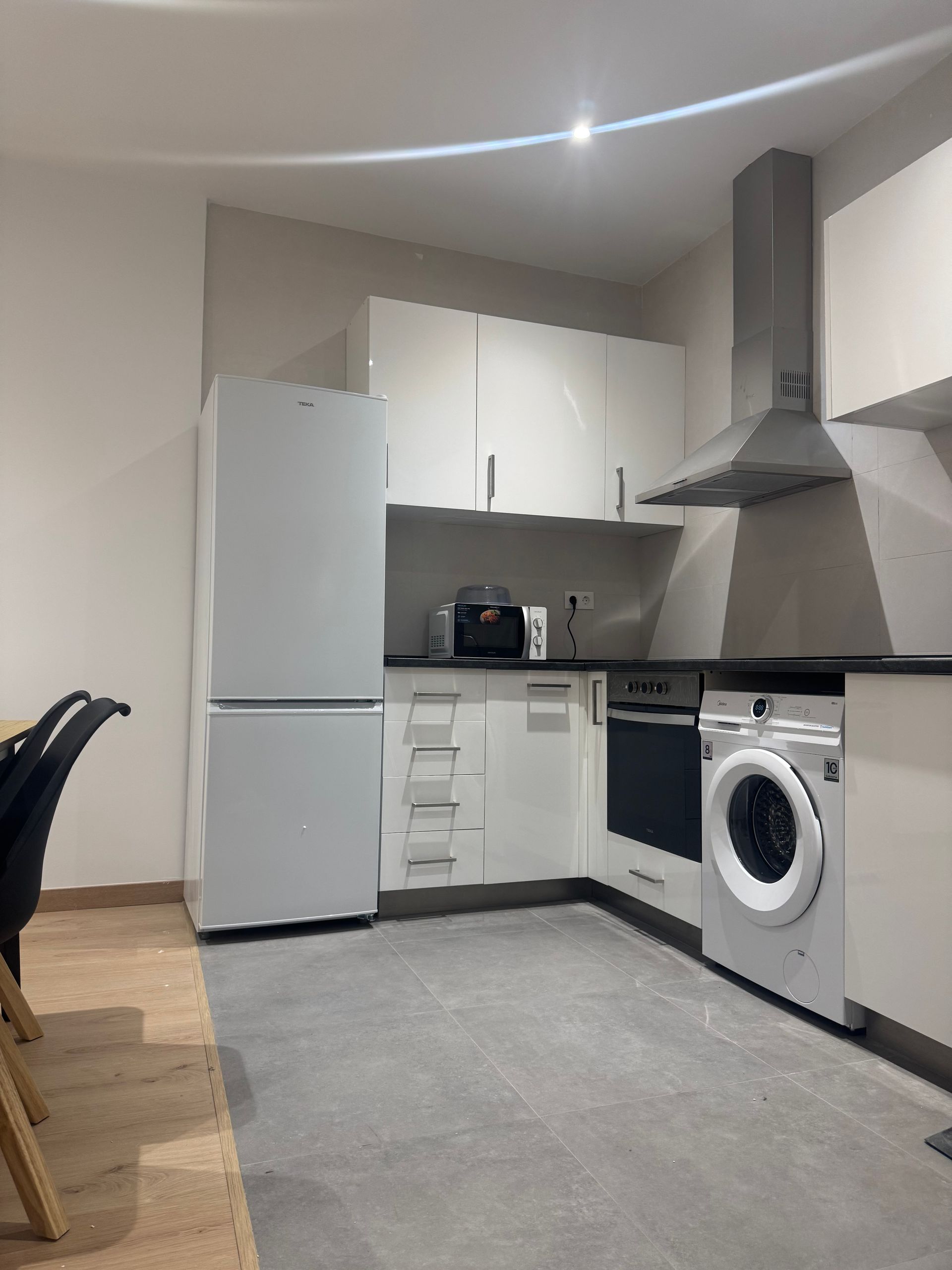 Cocina moderna y compacta con armarios blancos, nevera, horno, lavadora y suelo gris.