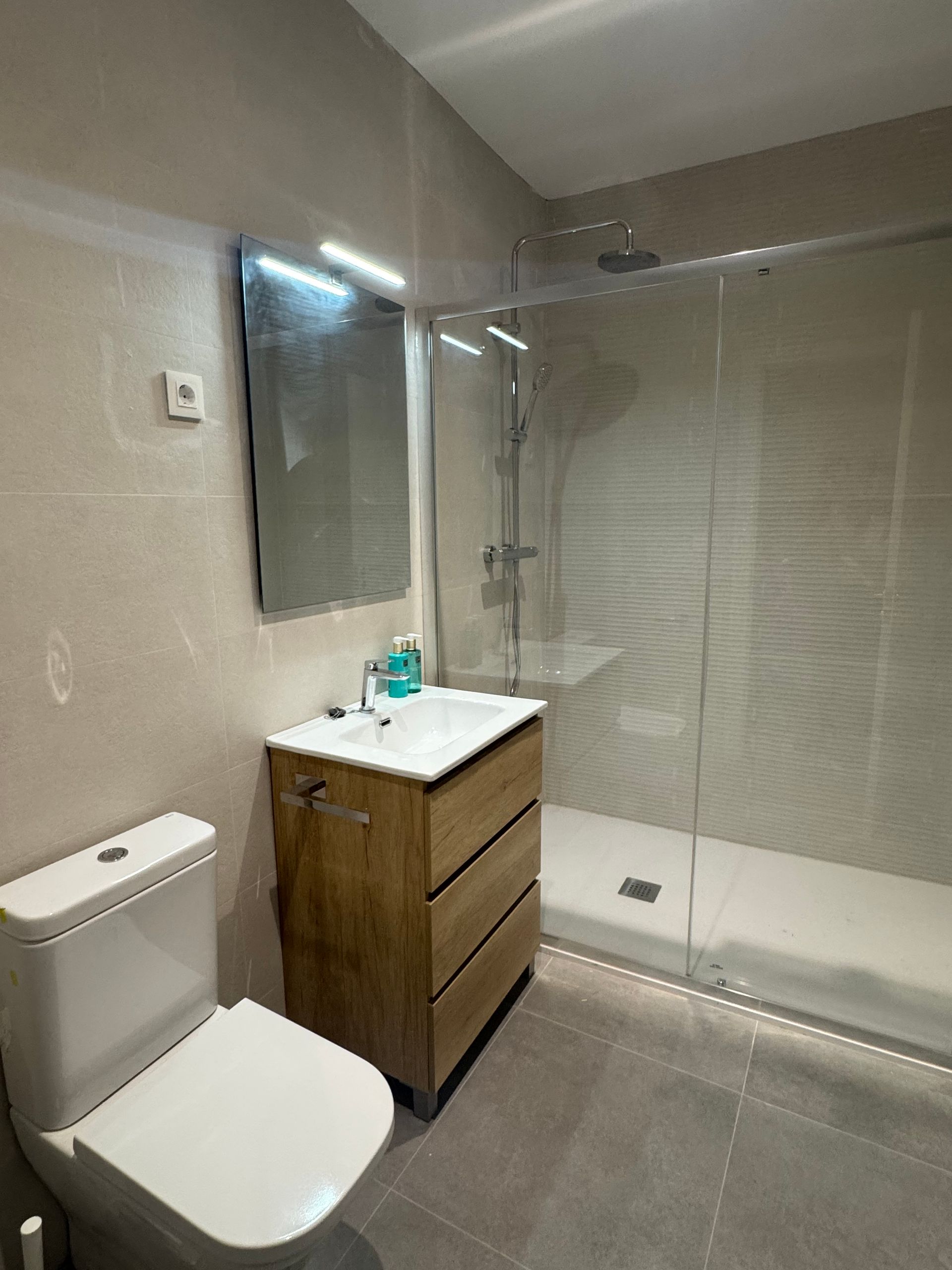 Baño moderno con inodoro, lavabo de madera, espejo y cabina de ducha de cristal.