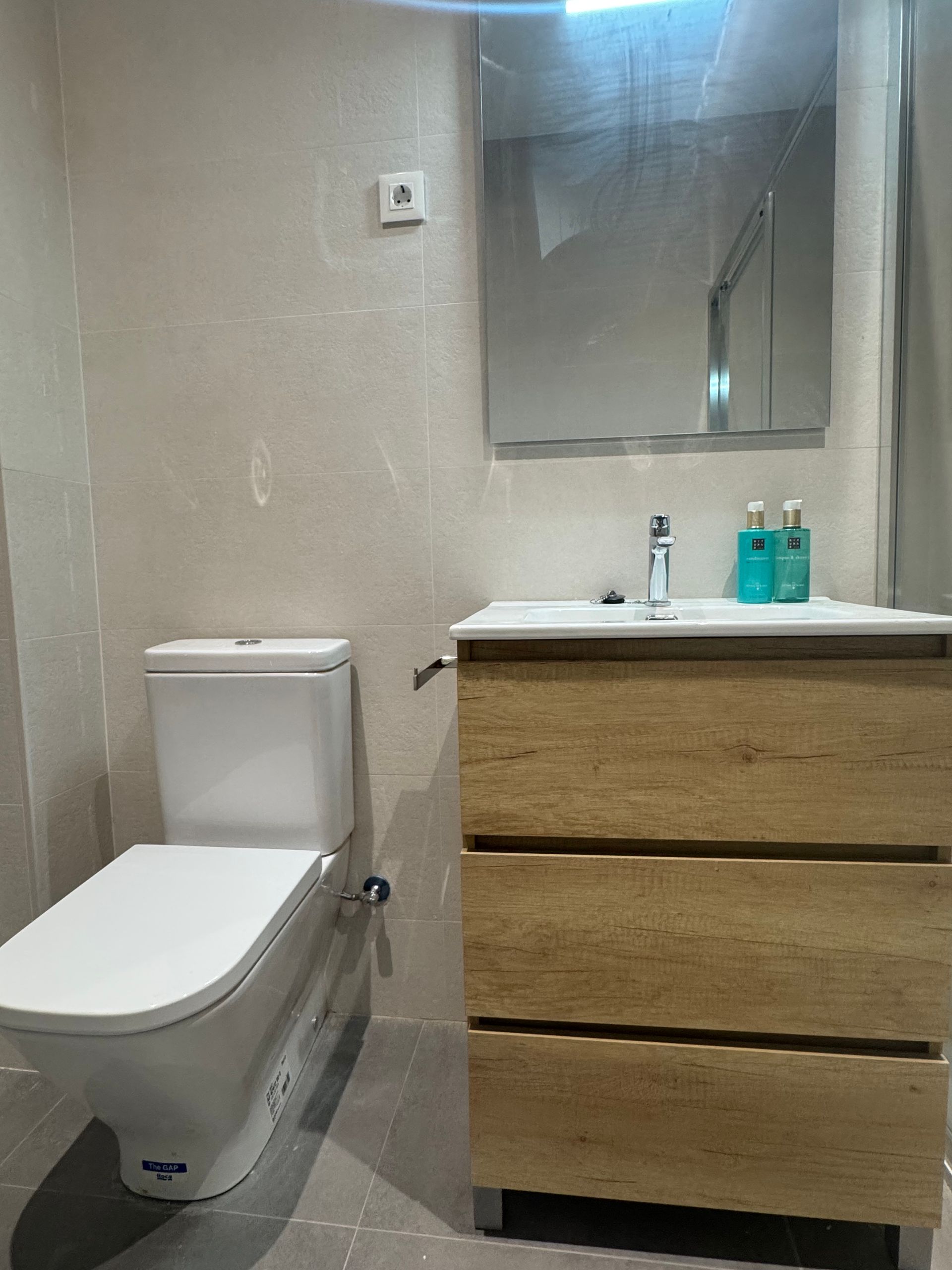 Baño pequeño con inodoro, tocador de madera, espejo, lavabo y artículos de aseo sobre la encimera.