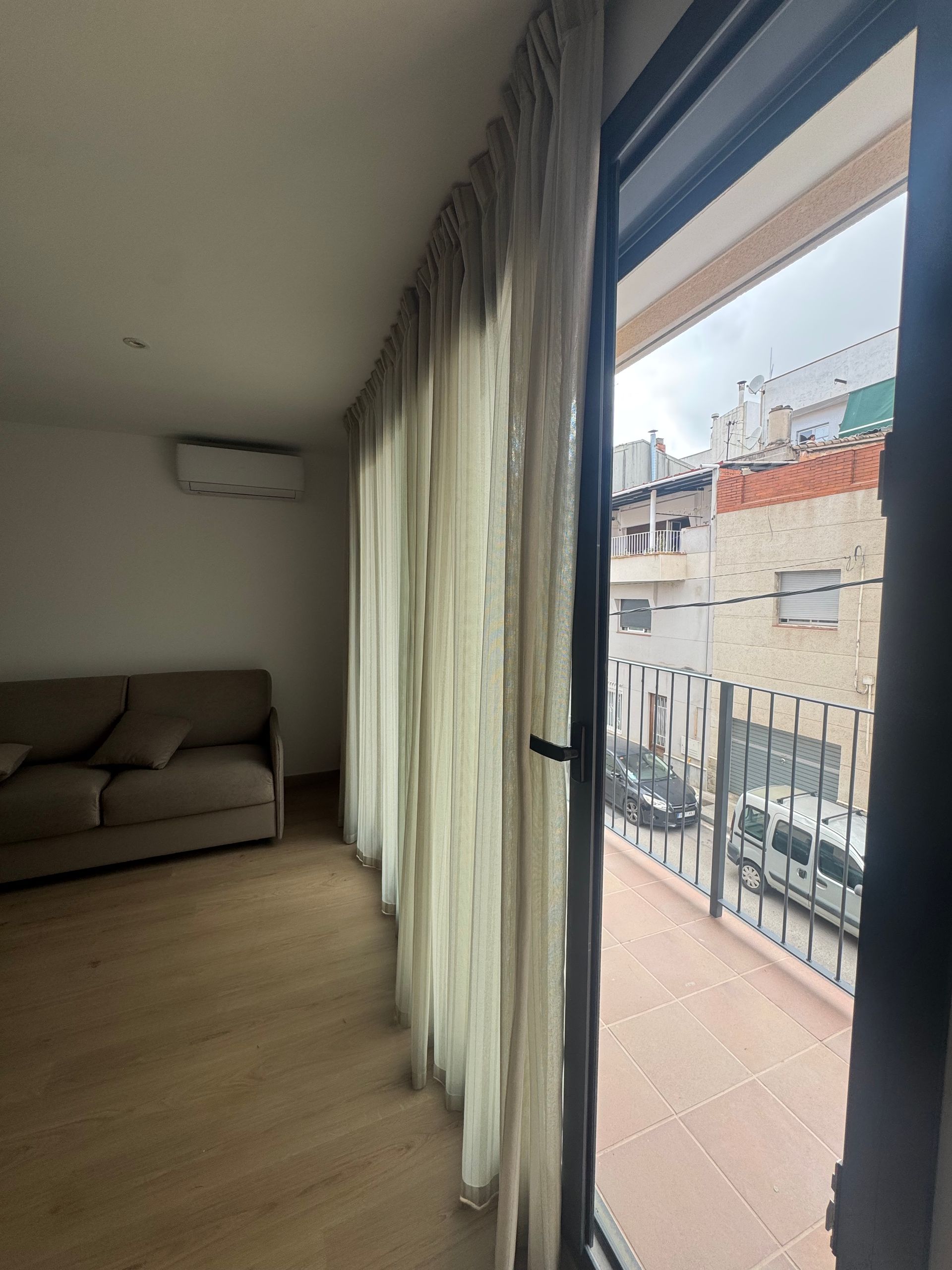 Sala de estar con sofá y cortinas transparentes junto a una puerta que da al balcón, con vistas luminosas a la ciudad.