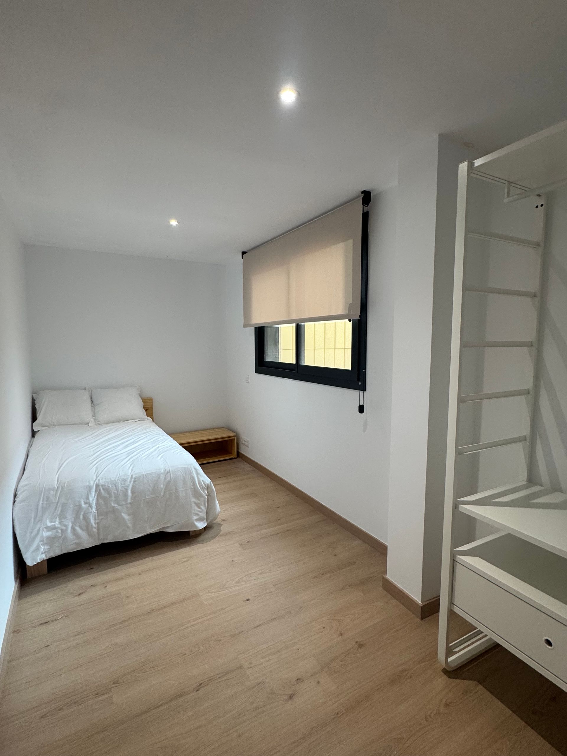 Habitación luminosa y minimalista con cama blanca, suelo de madera y ventana con persiana beige.