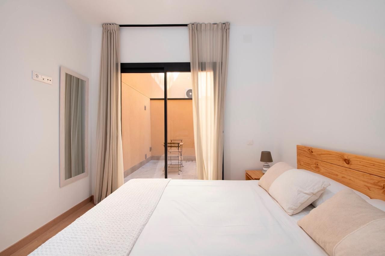 Habitación luminosa con cama blanca, cabecero de madera y puertas correderas que dan a un balcón o patio.