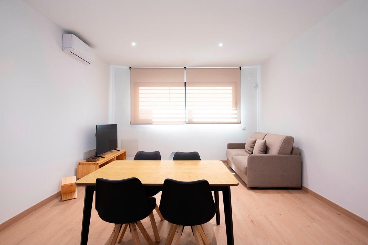 Sala de estar minimalista con sofá, mueble para TV, mesa de comedor y dos sillas negras junto a una ventana.