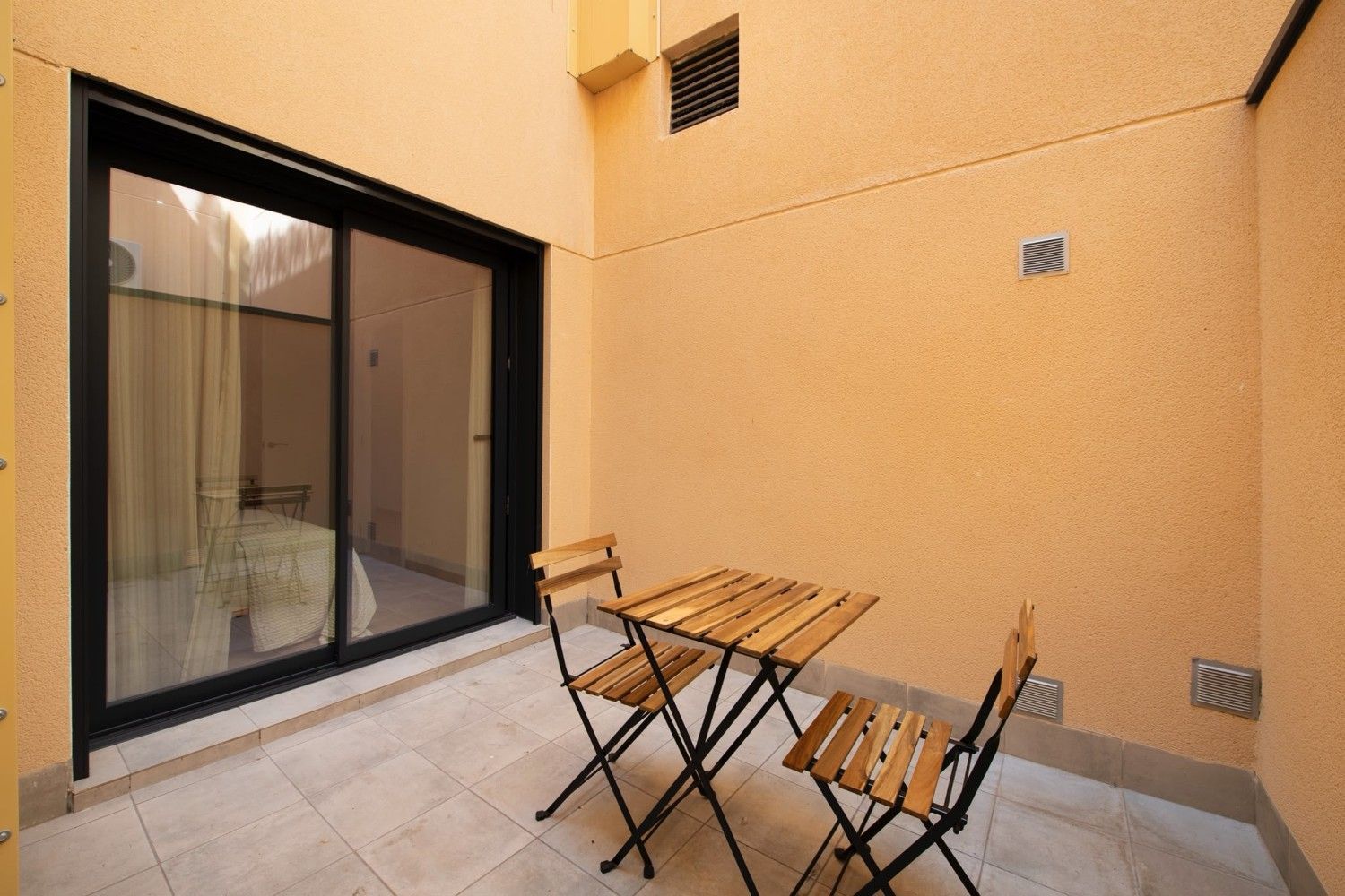 Pequeño patio con mesa y sillas de madera junto a una puerta corrediza de cristal y paredes de color beige.
