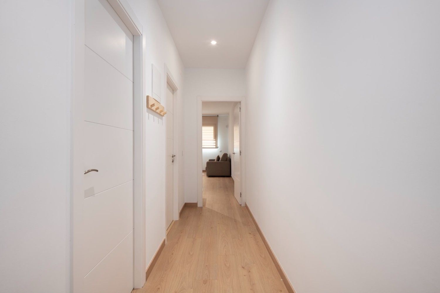 Pasillo luminoso con paredes blancas, suelo de madera clara, puertas a la izquierda y una habitación visible al final.