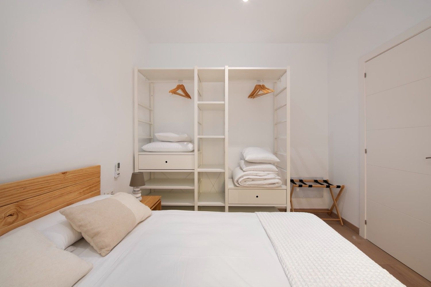 Dormitorio blanco luminoso con una cama en primer plano y un armario abierto con sábanas dobladas y perchas contra la pared.