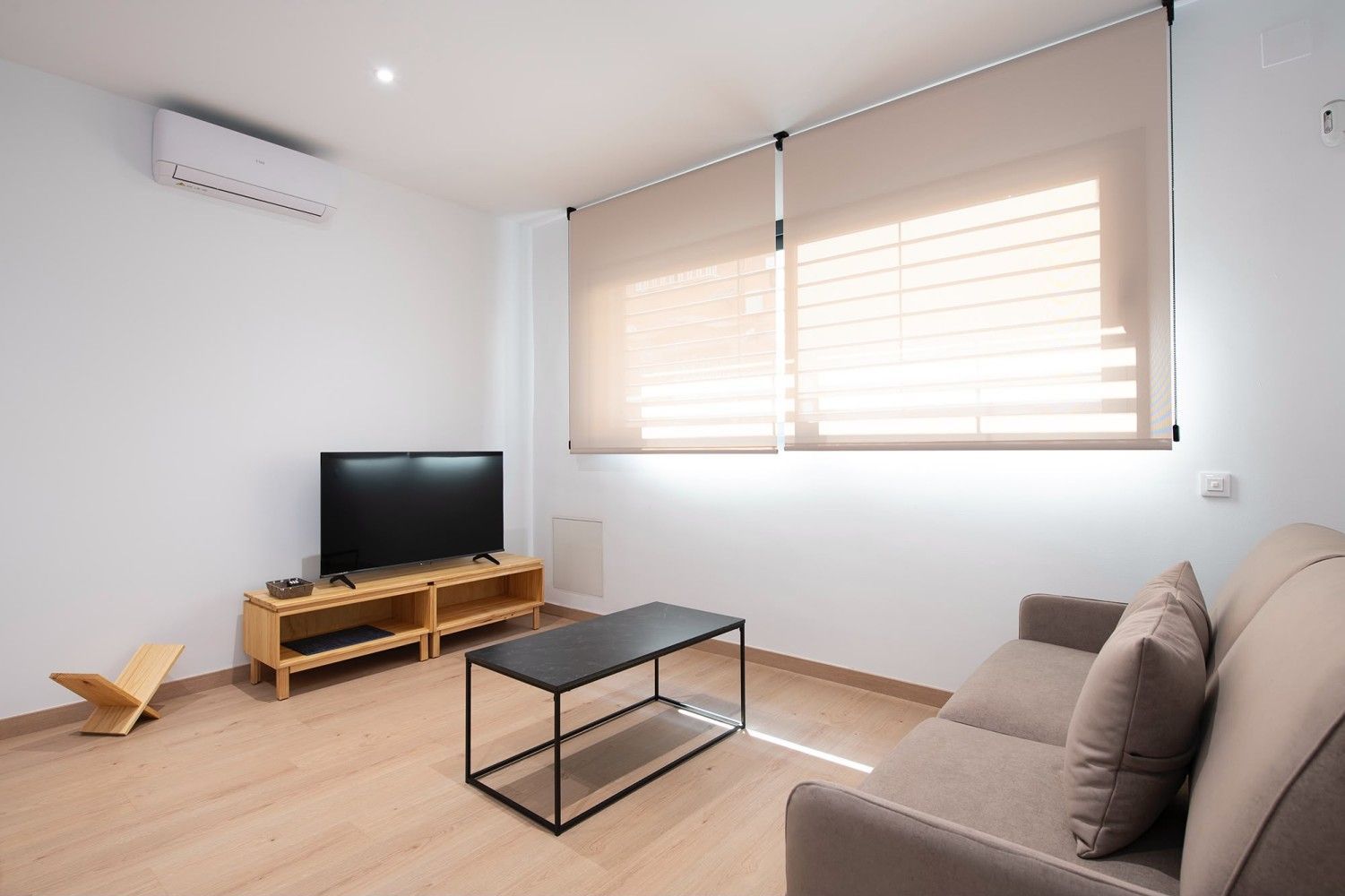 Luminoso salón con sofá, televisor, mesa de centro y grandes persianas.
