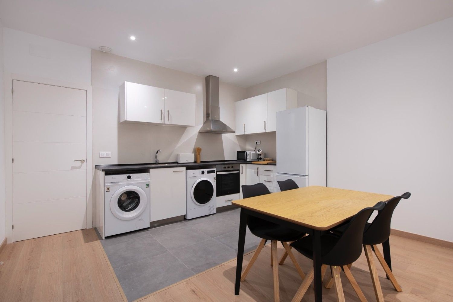 Cocina y comedor modernos y luminosos con armarios blancos, electrodomésticos y una mesa de madera con sillas negras.