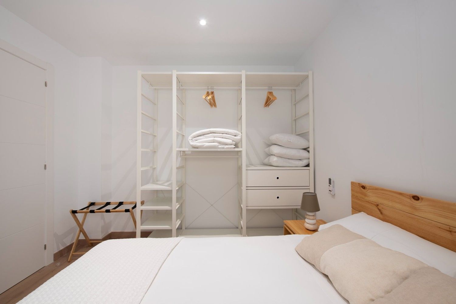 Dormitorio minimalista blanco con armario abierto, estructura de cama de madera y sábanas dobladas.