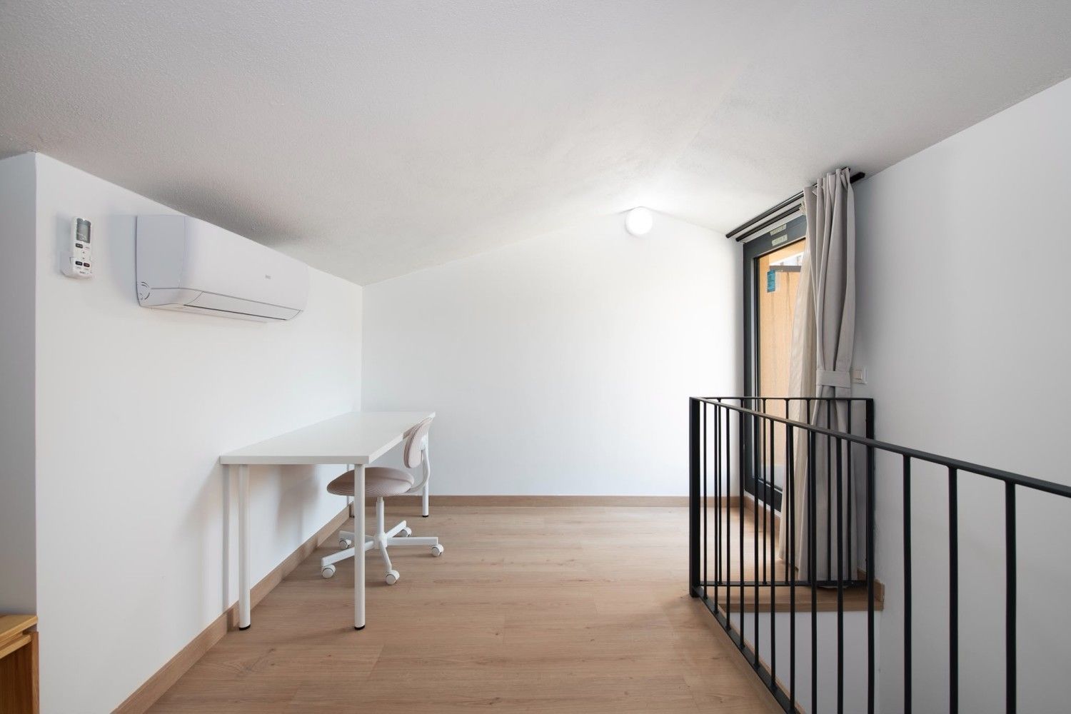 Despacho tipo loft minimalista con escritorio, silla, aire acondicionado, suelo de madera y luz natural.