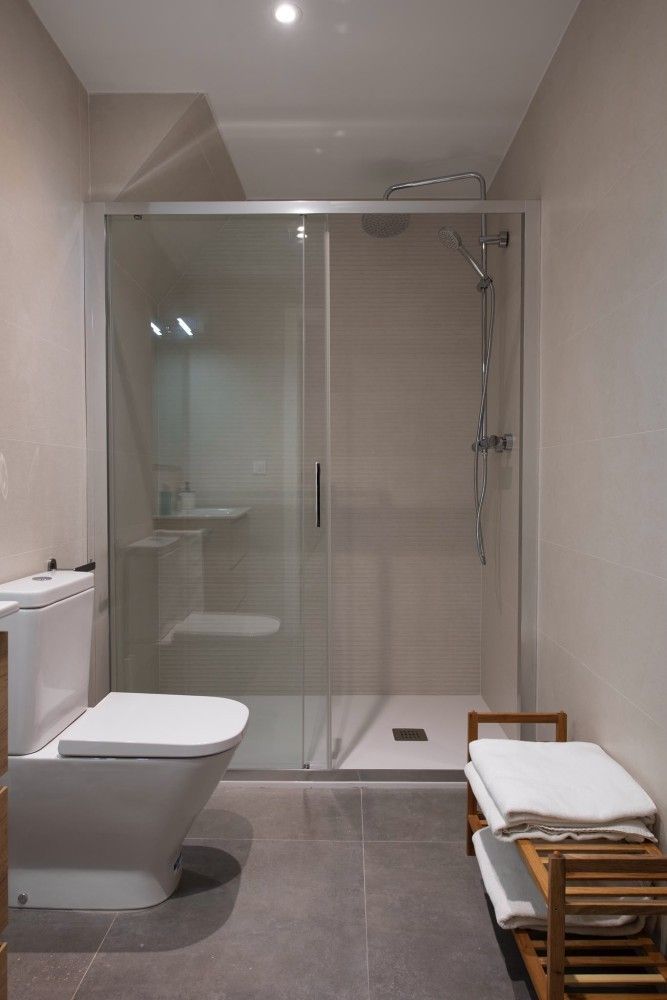 Baño moderno con ducha de cristal, inodoro y banco de madera sobre suelo gris.