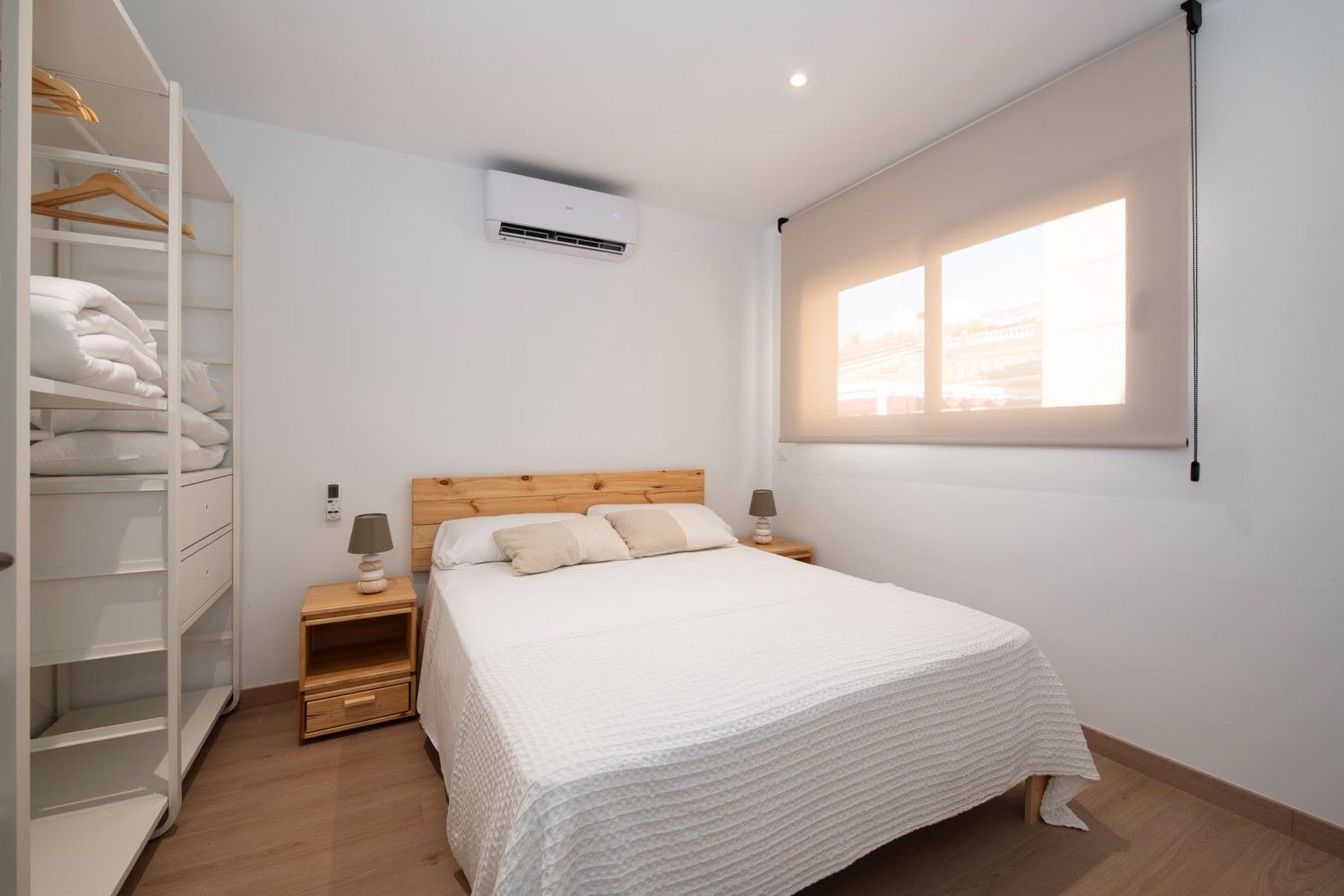 Habitación luminosa con cama, mesitas de noche, estanterías, aire acondicionado y ventana con persianas.