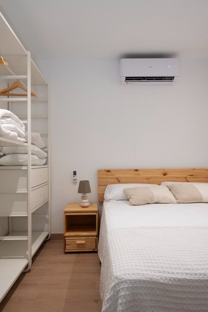 Dormitorio minimalista con cama blanca, mesita de noche de madera, estante y aire acondicionado mural.