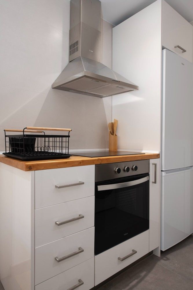 Cocina compacta de color blanco con encimeras de madera, horno, campana extractora y frigorífico alto.