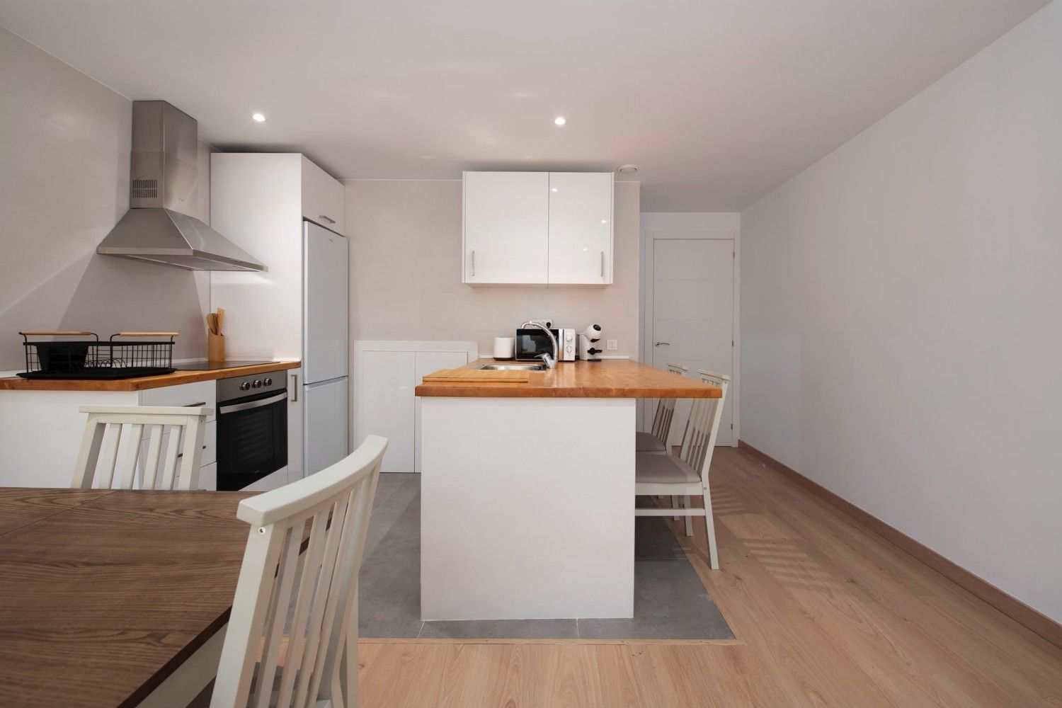 Cocina moderna y luminosa con armarios blancos, encimeras de madera y una isla central en un espacio diáfano.