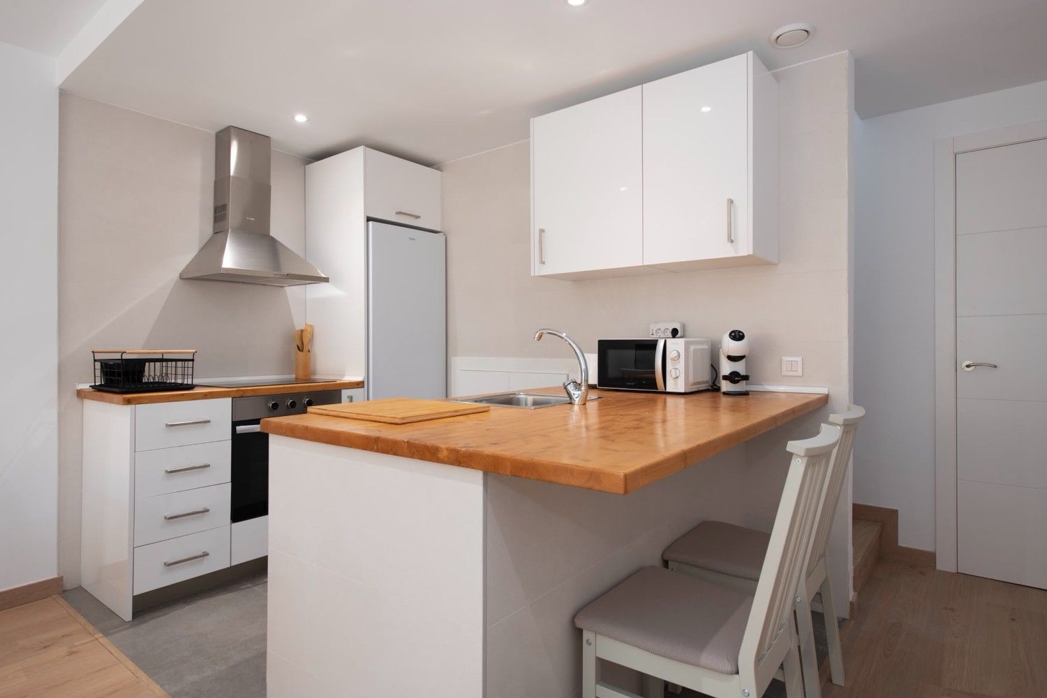 Cocina moderna blanca con isla de madera, taburete de bar, campana extractora de acero inoxidable y electrodomésticos.