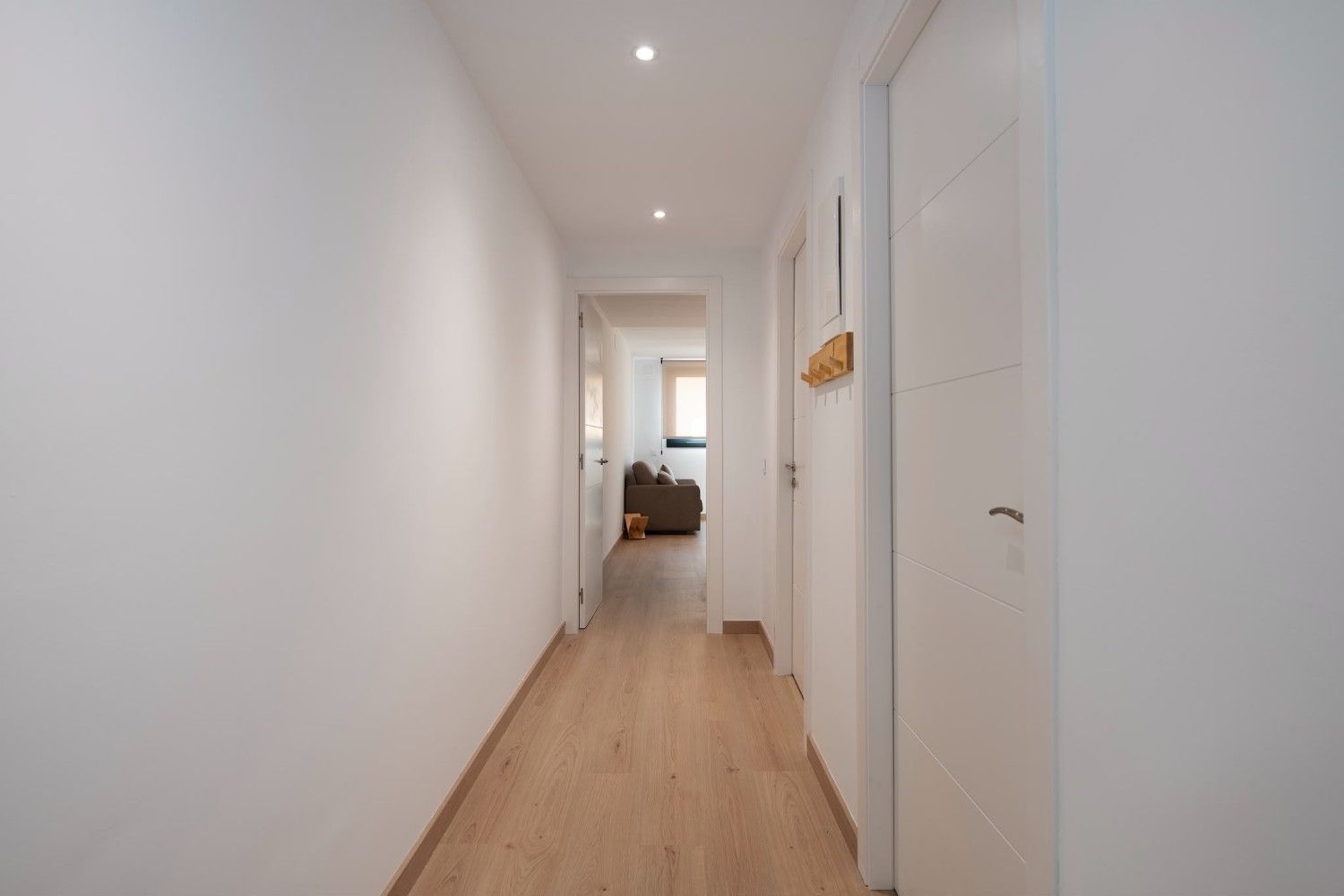 Pasillo luminoso con paredes blancas, suelo de madera y un dormitorio visible al final.