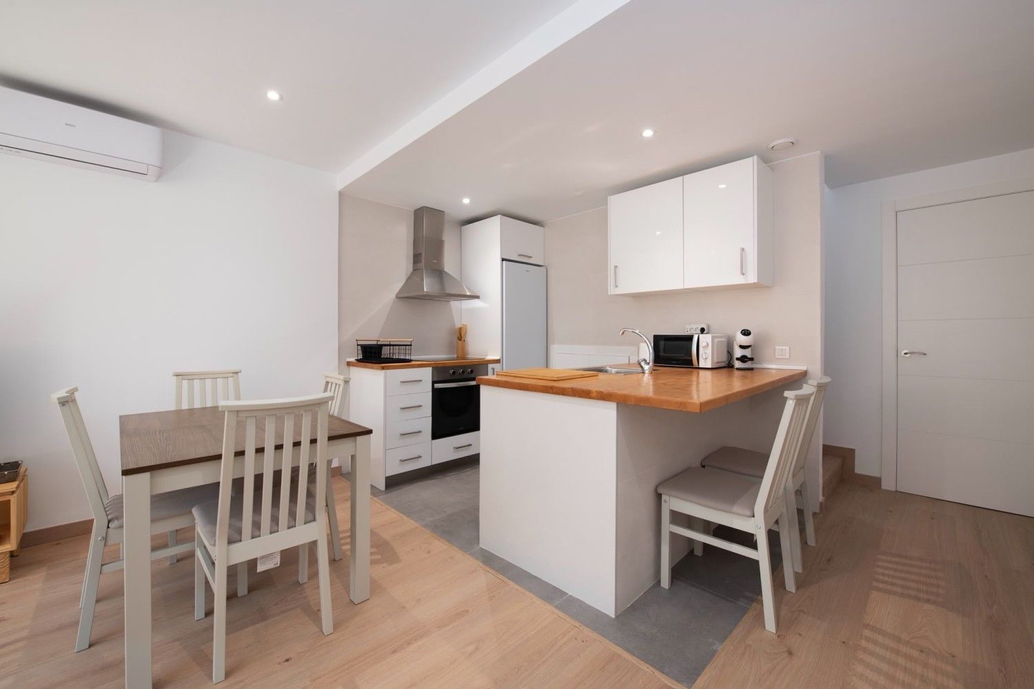 Cocina-comedor moderna y luminosa con armarios blancos, suelos de madera y una isla con sillas.