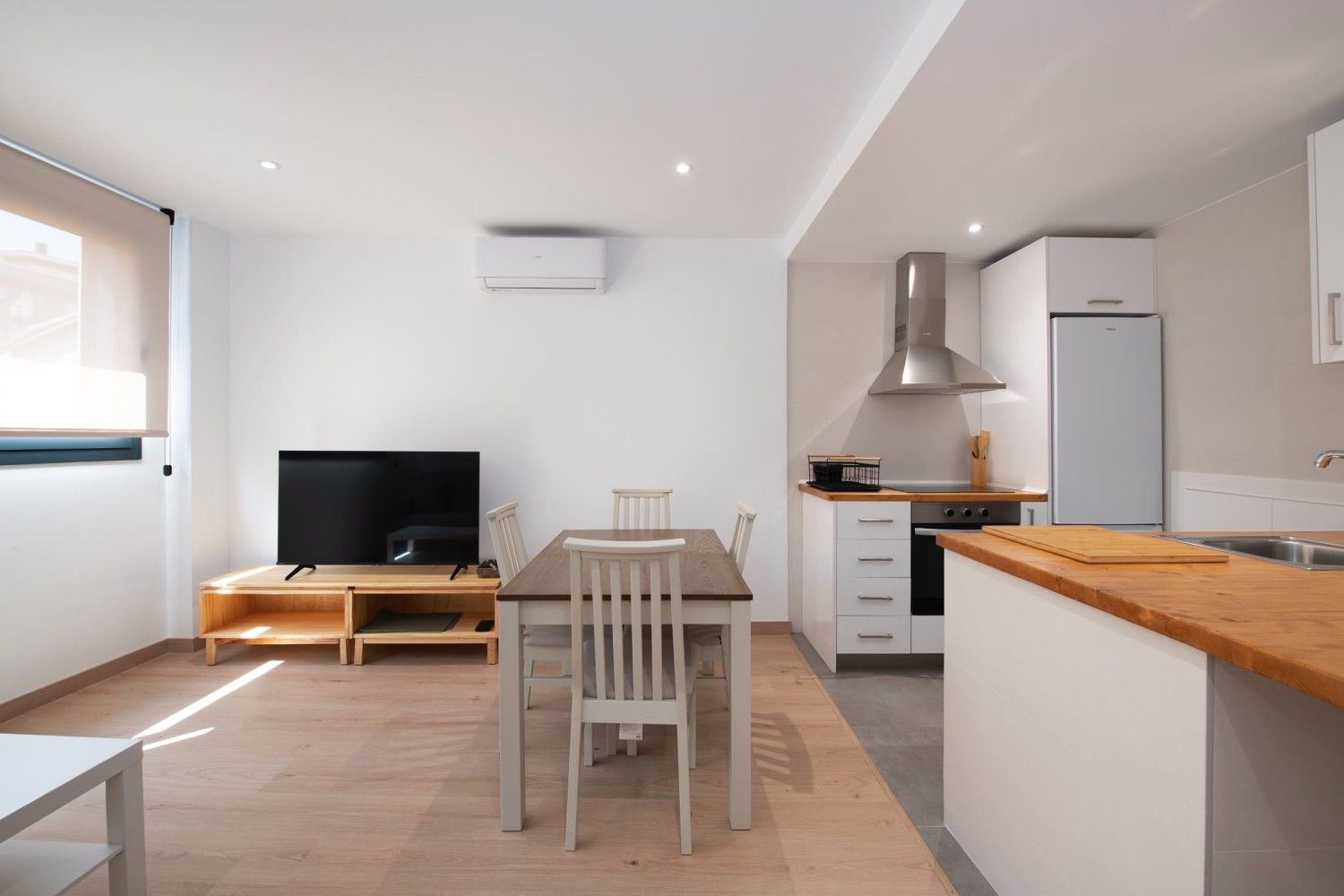 Cocina y sala de estar de planta abierta con paredes blancas, suelo de madera y pequeña mesa de comedor.