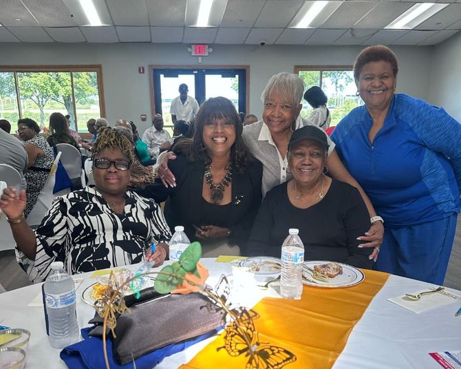 Five smiling people pose at a banquet table in a bright community hall, with a yellow tablecloth and drinks.