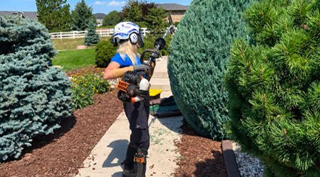 Worker using a hedge trimmer beside tall shrubs in a landscaped garden path.
