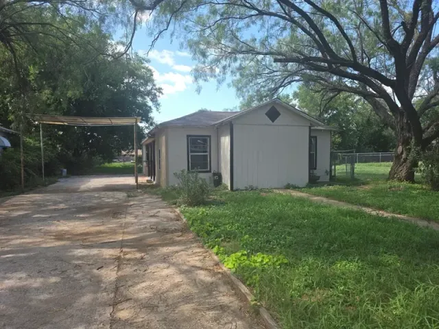 Small white house with a carport, driveway, and green yard under large trees
