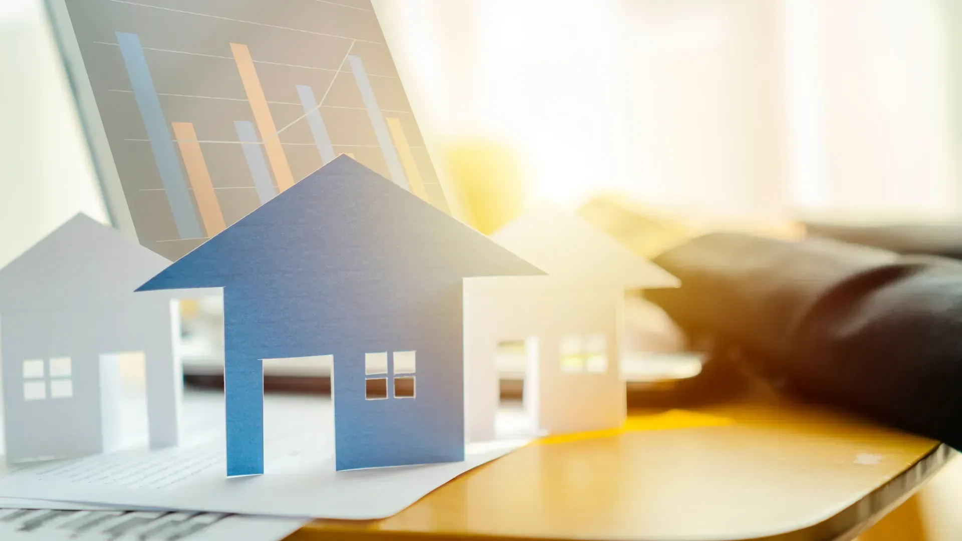 Paper house models on a desk with sunlight and financial charts in the background