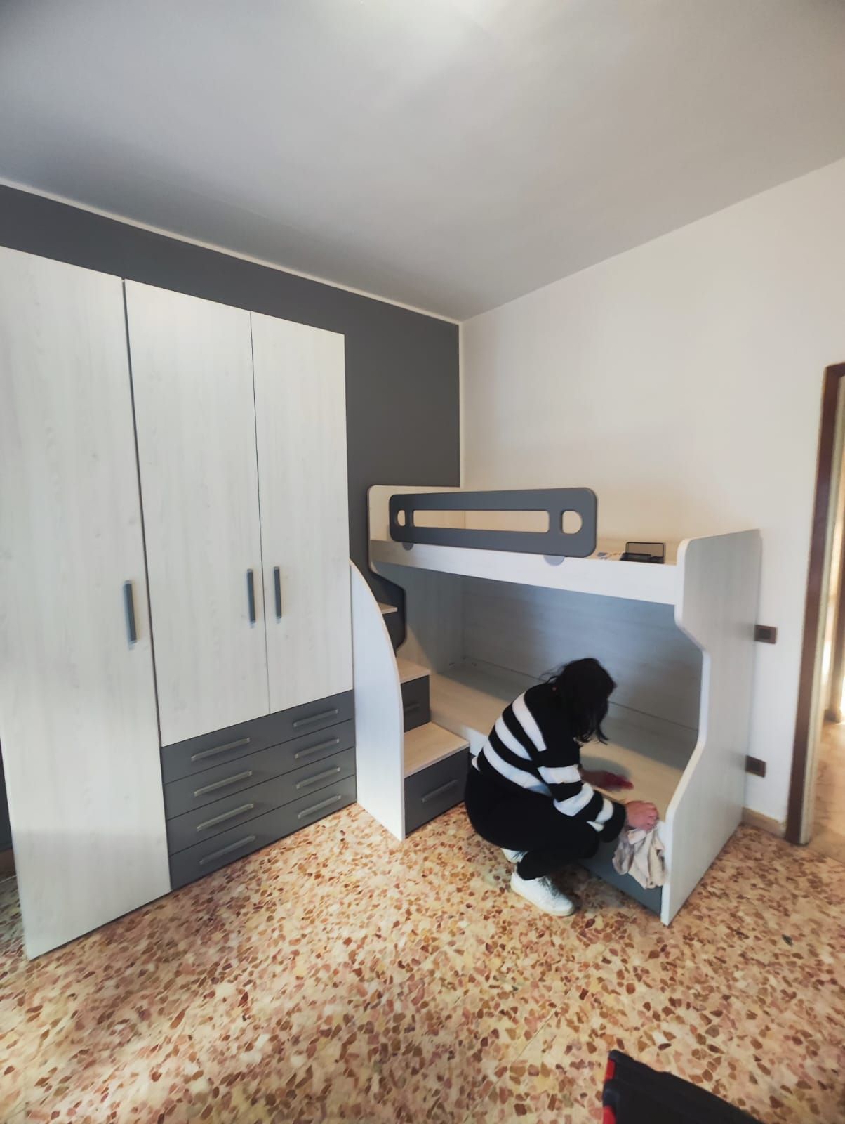 Una persona è accovacciata accanto a un letto a castello in una camera da letto, vicino a un armadio bianco con maniglie nere.