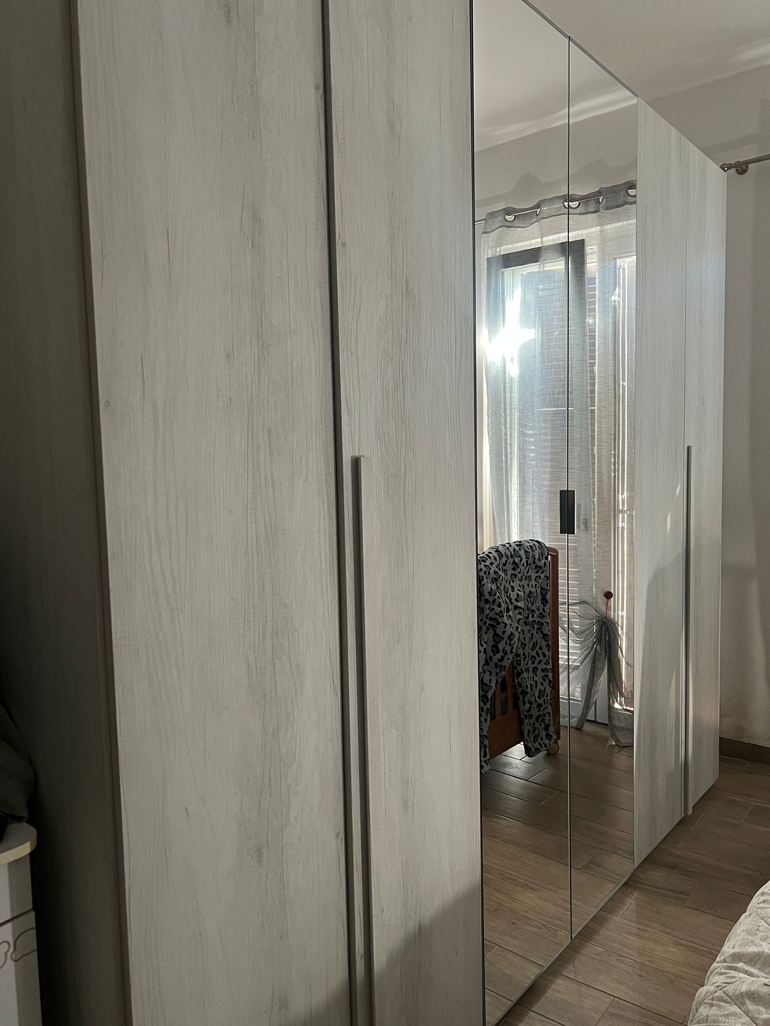 Un alto armadio bianco con ante a specchio in una stanza stretta, che riflette gli abiti e la porta.