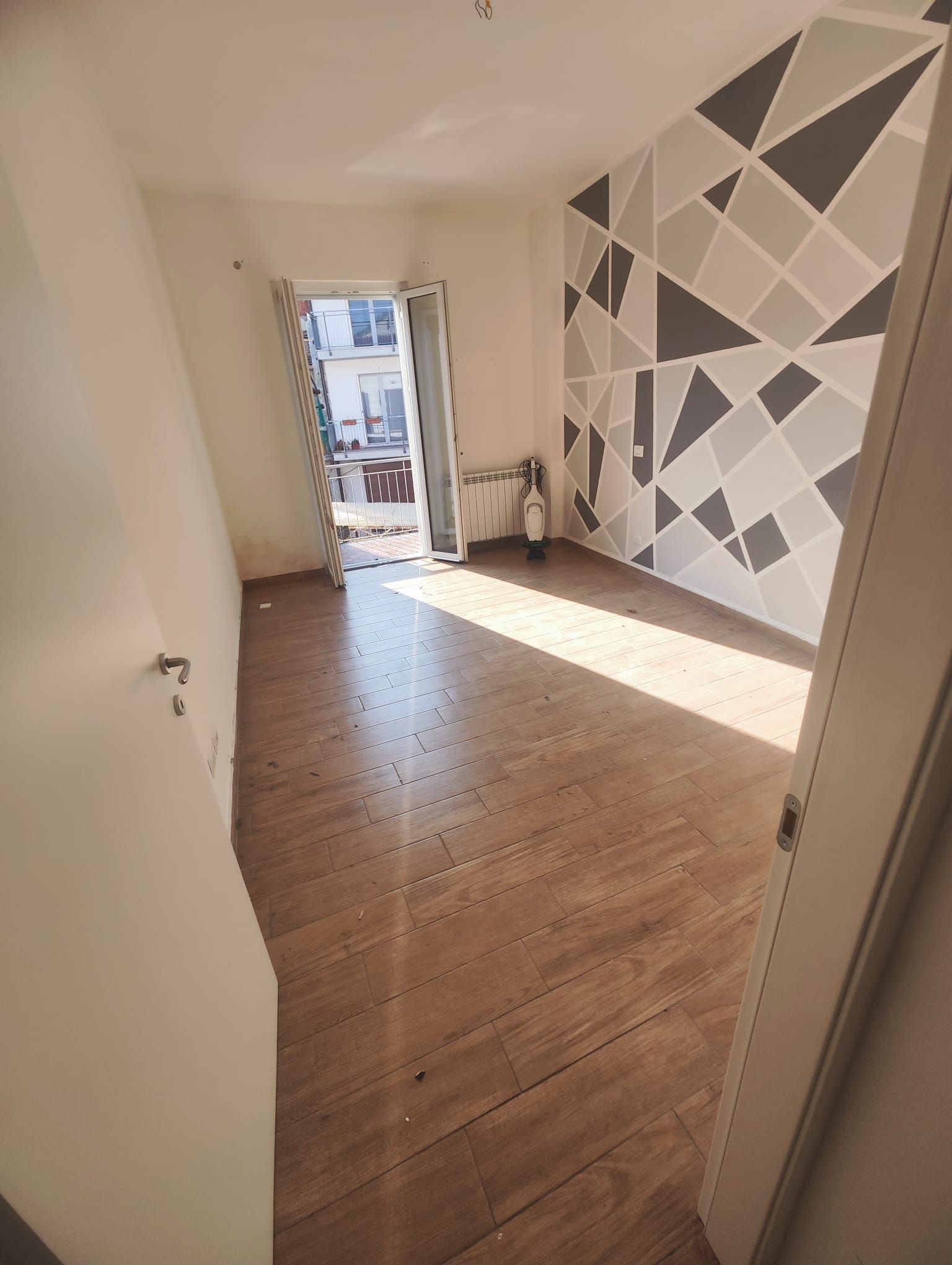 Stanza stretta e vuota con pavimento in legno e parete grigia geometrica, illuminata dalla luce del sole che filtra da una porta in fondo.