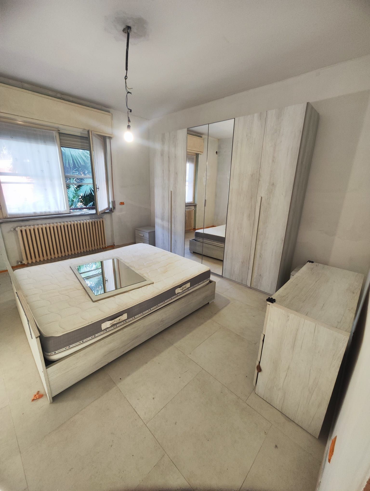 Camera da letto vuota con letto, armadio, cassettiera, finestra e lampadario a sospensione.
