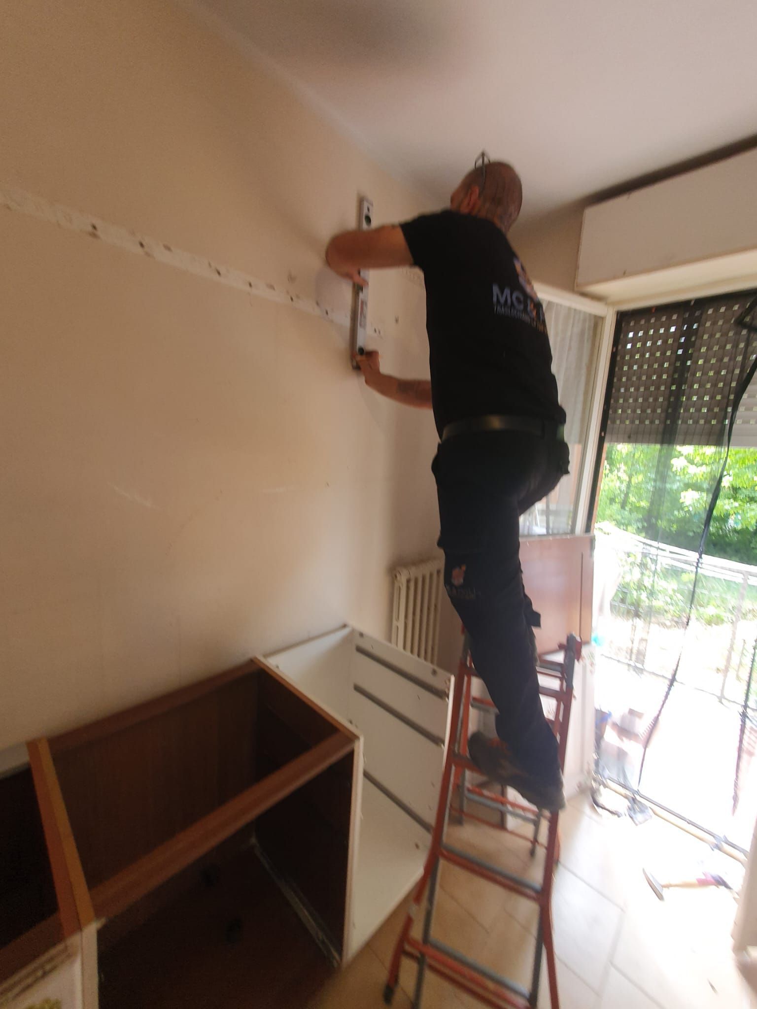 Una persona in piedi su una scala sta lavorando su una parete in una stanza con una scrivania e una porta-finestra che dà sul balcone.