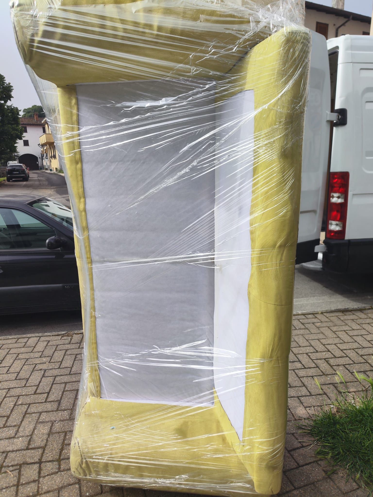 Un grande oggetto imbottito avvolto in giallo si trova su un marciapiede, avvolto nella plastica accanto a veicoli parcheggiati.
