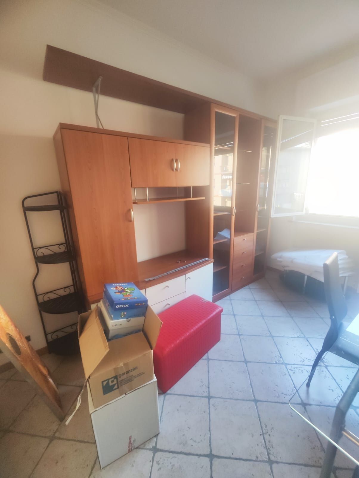 Stanza ingombro di oggetti con un mobile porta TV in legno, scatole, un pouf rosso e luce del sole che filtra da una finestra luminosa.