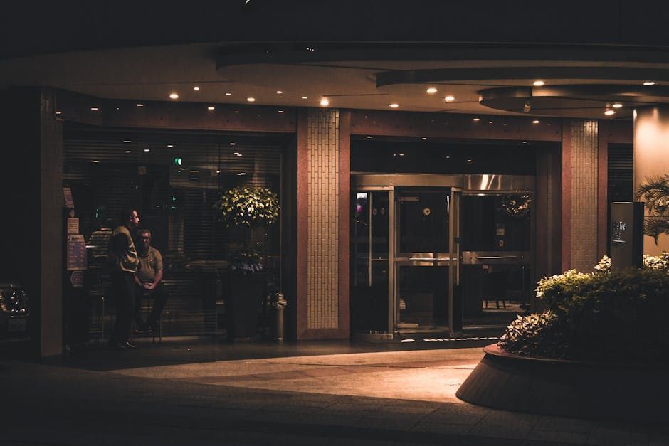 Hotel entrance at night, glass doors, lit interior, people visible, small planters.
