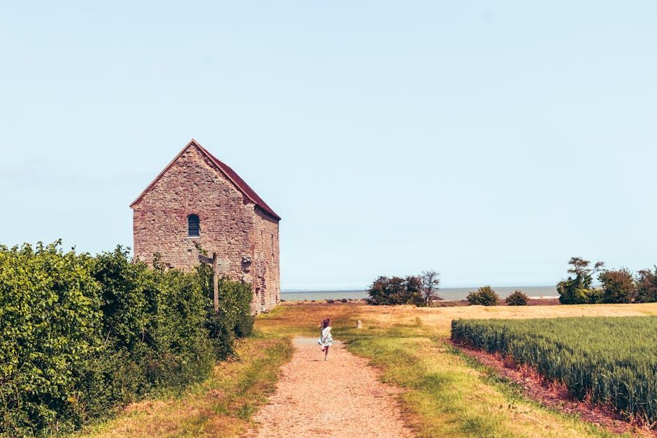 A person walks down a dirt path towards a stone building near a field and the sea under a blue sky.