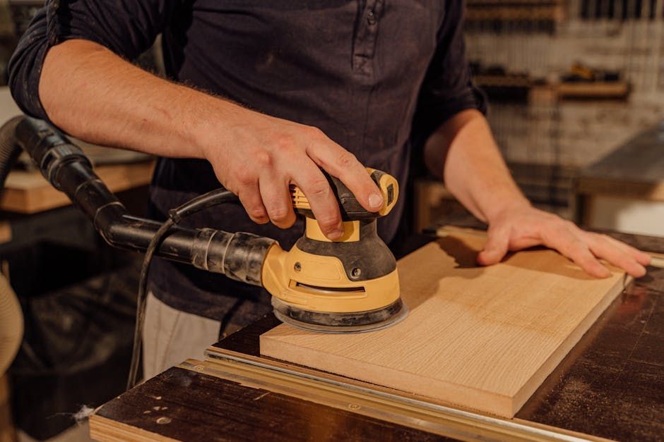 Person sanding a wood plank with a yellow power sander, workbench in the background.