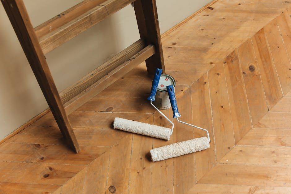 Wooden ladder leaning against a wall, two paint rollers on a herringbone wood floor.