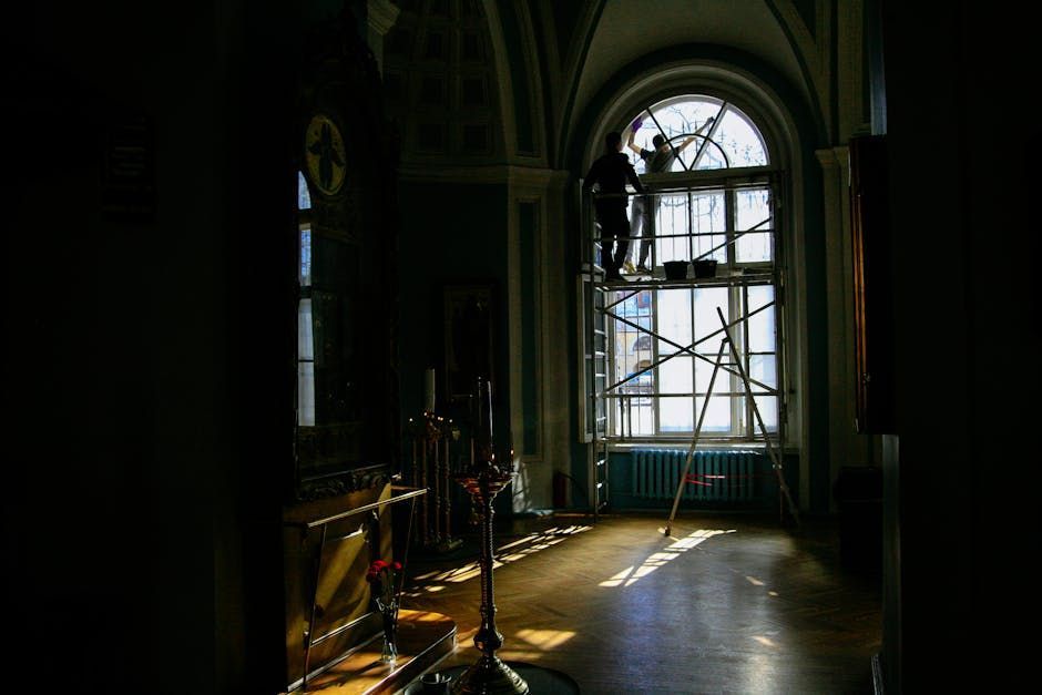 Two people on scaffolding cleaning a large arched window in a dimly lit room.