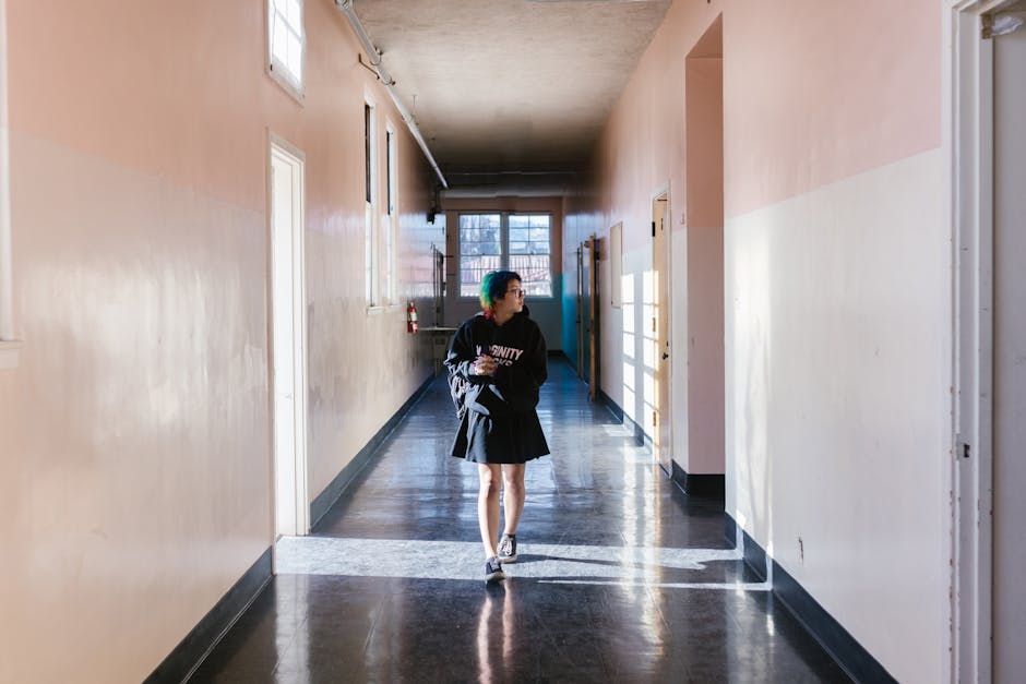 Person walking down a long hallway with pink and white walls, holding books. Sunlight streams in.