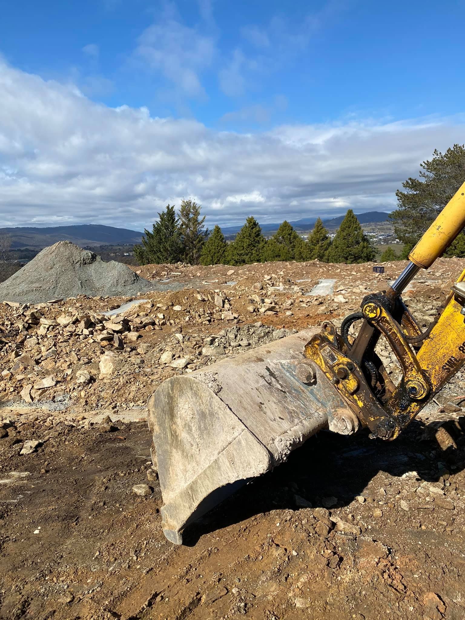 Excavator bucket scoops up rocks on a construction site with a mountain view and blue sky. — Brucic Construction in Dickson, ACT