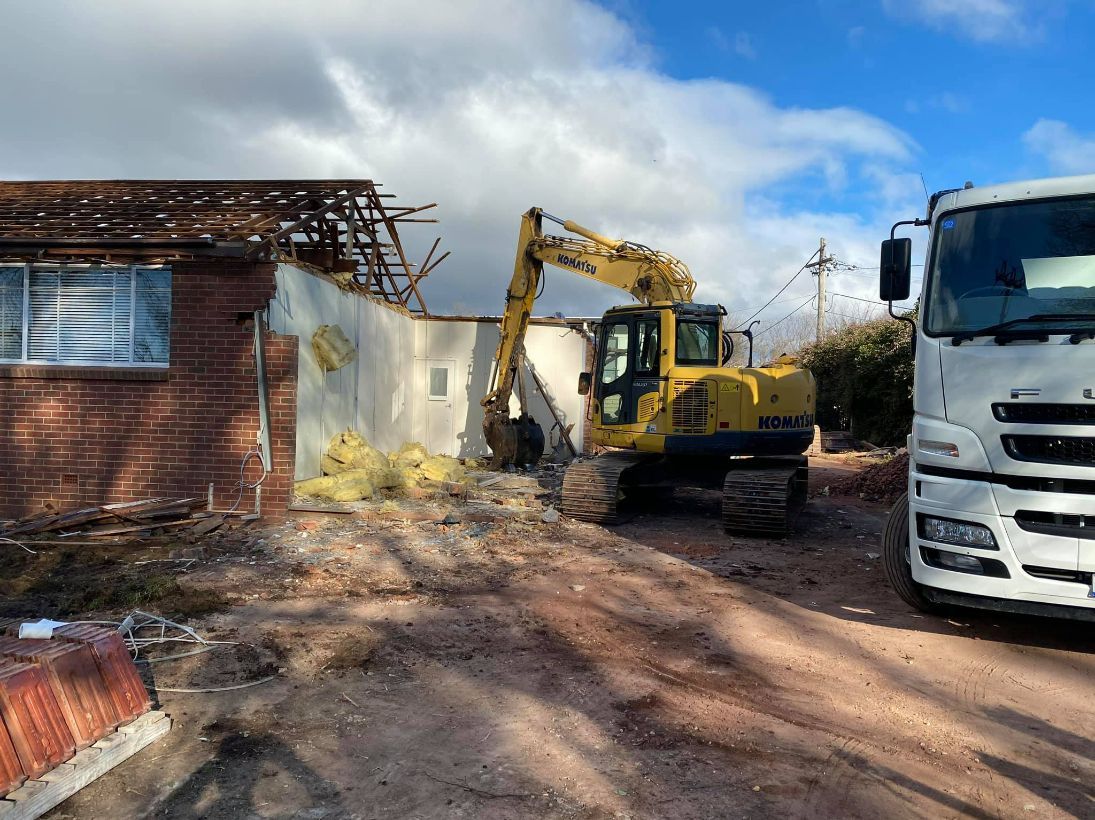 A yellow excavator demolishes a brick building, with a truck parked nearby on a dirt lot. — Brucic Construction in Queanbeyan, NSW