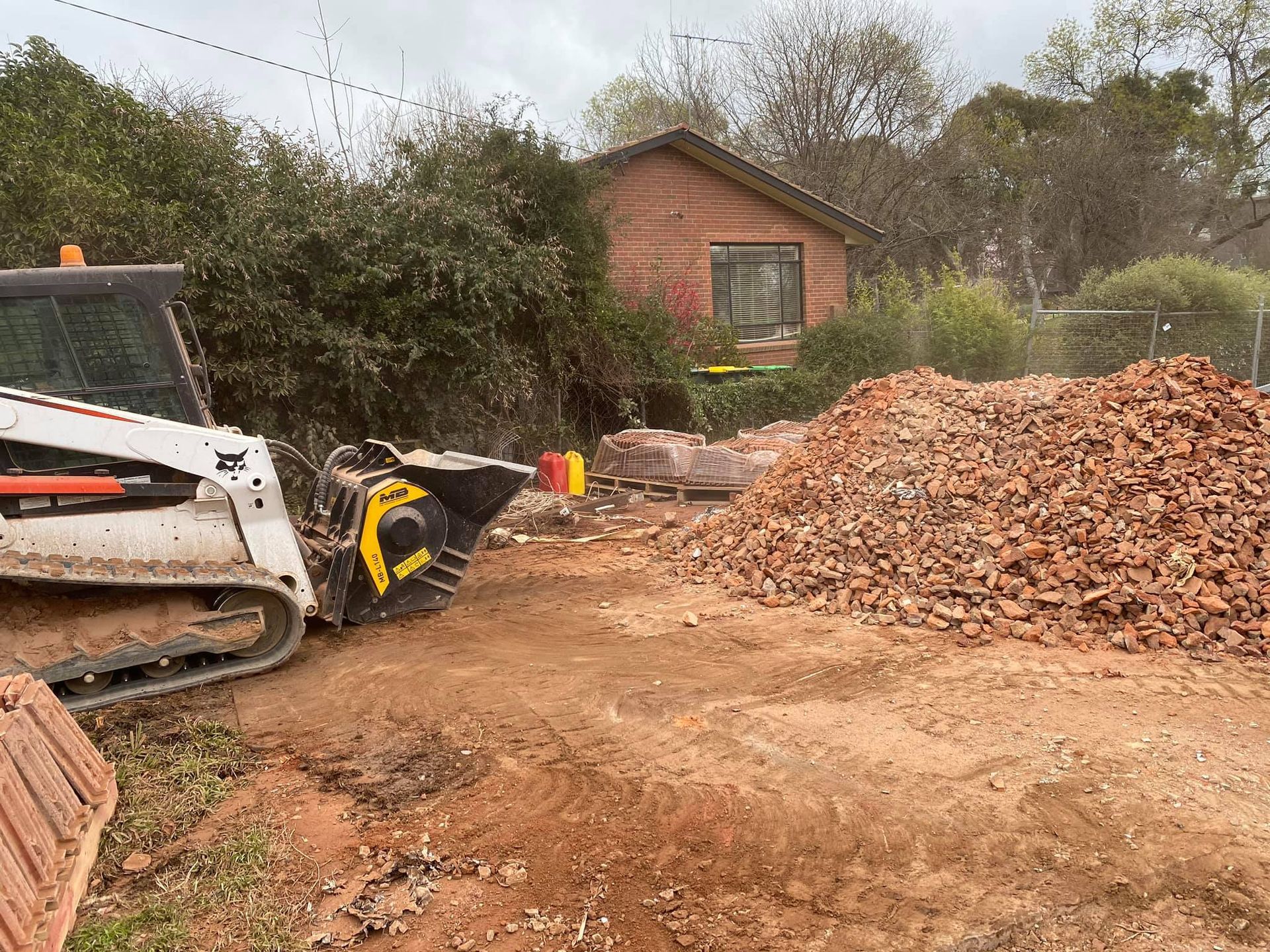 Bobcat with a grinder mulching brush, pile of debris in yard, brick building in background. — Brucic Construction in Dickson, ACT