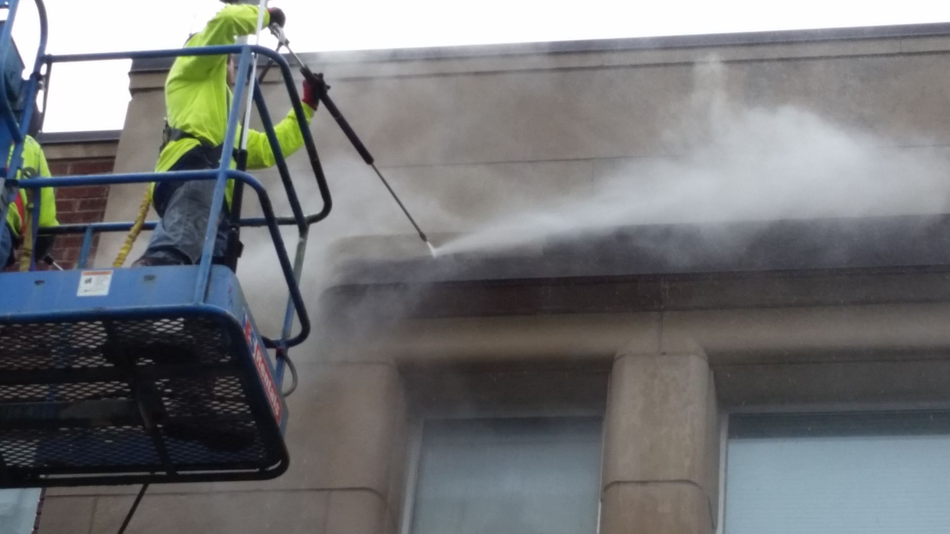 Two men are cleaning a building with a high pressure washer.