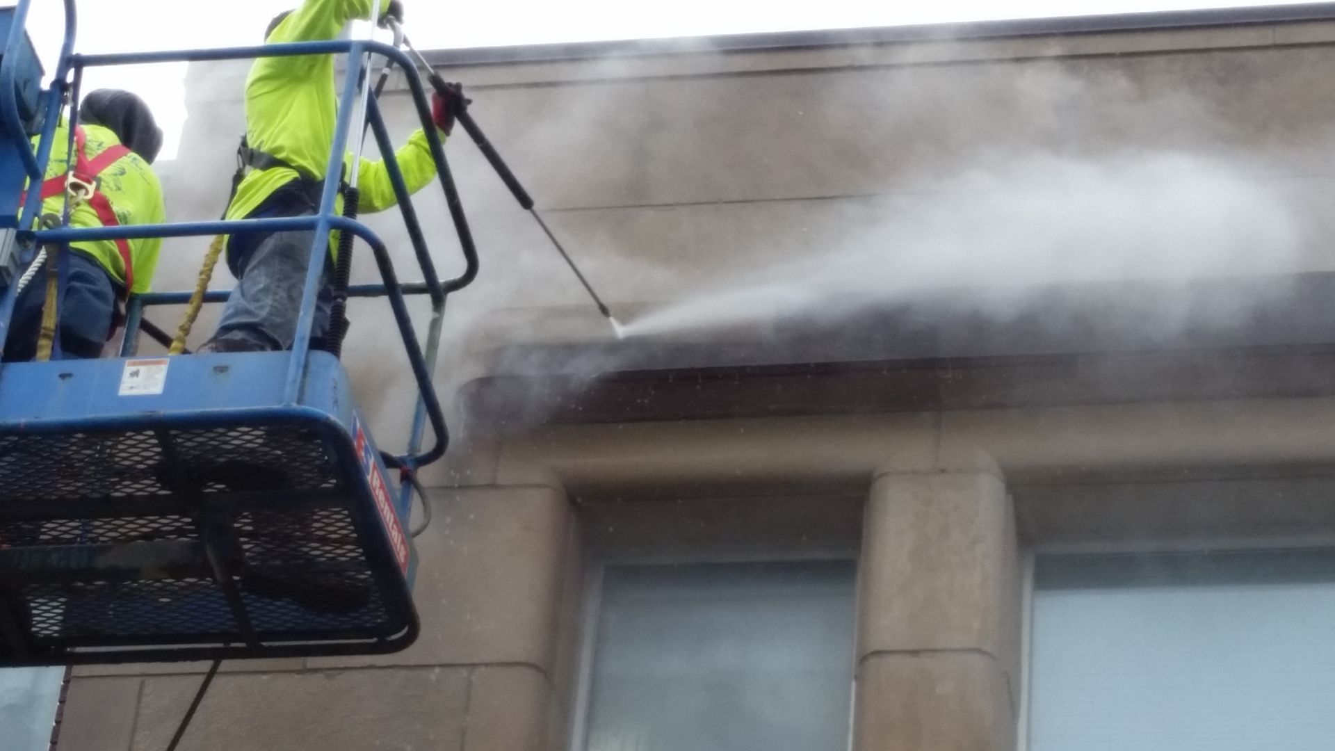 Two men are cleaning a building with a high pressure washer.