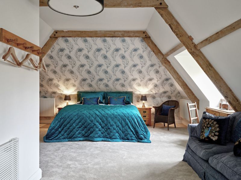 Bright attic bedroom with teal bedspread, exposed beams, patterned wallpaper, and a gray sofa